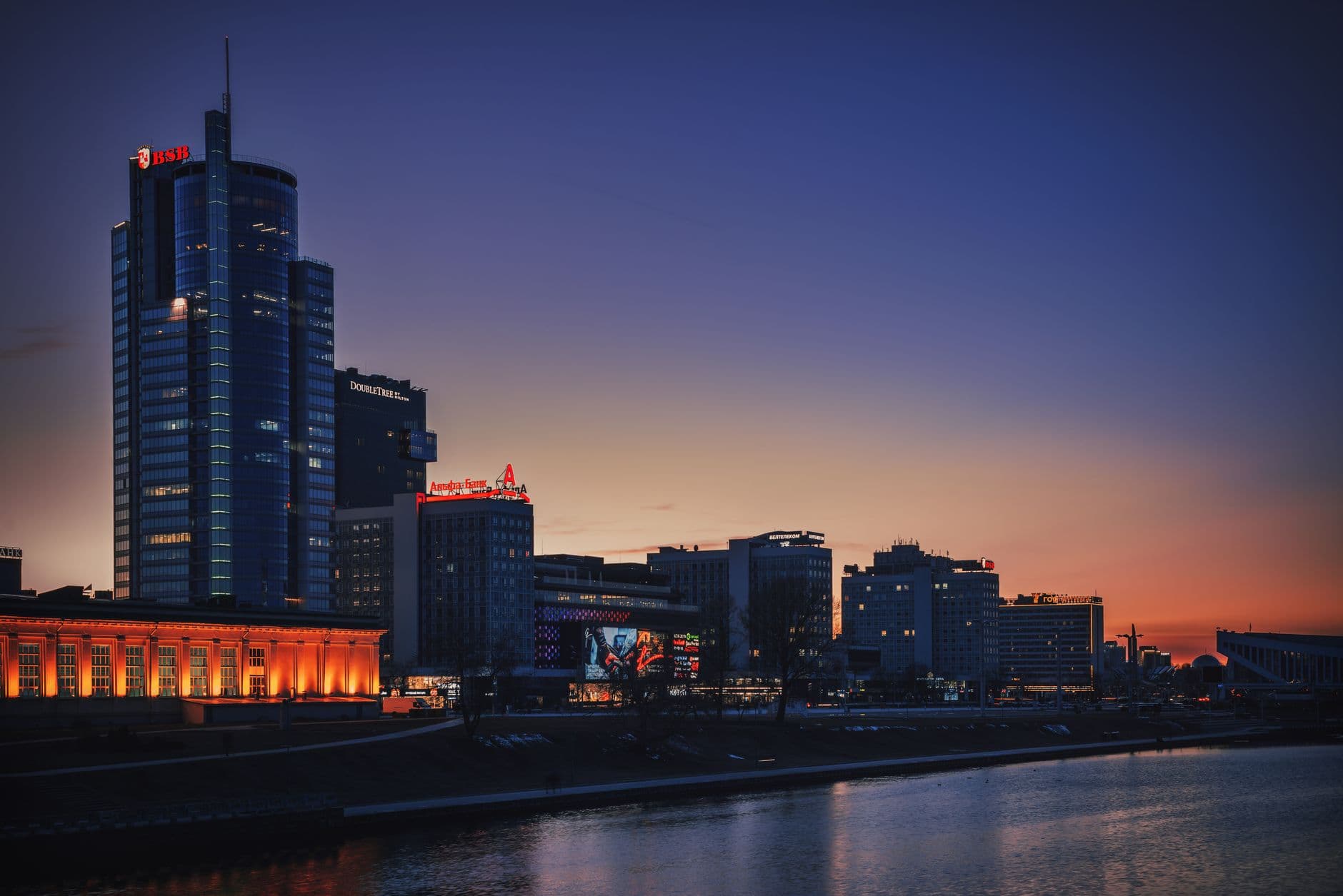 A vibrant night view of Minsk city center, with illuminated buildings and modern architecture reflecting in a wet street, suggesting a dynamic urban travel experience.