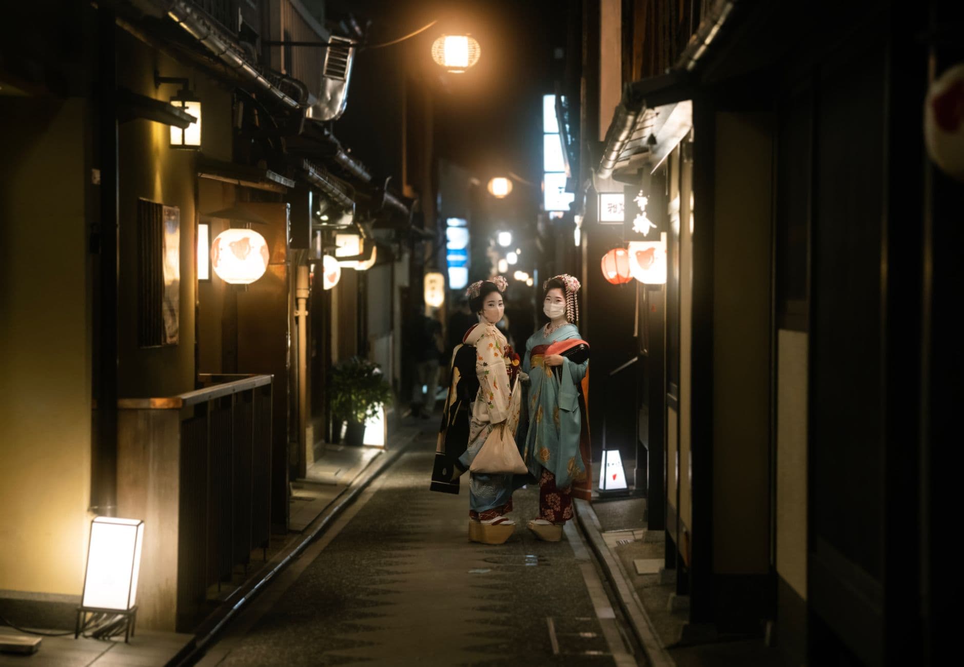 A narrow, historic street in Kyoto's Gion district at night, illuminated by traditional lanterns, with a woman walking away from the camera.