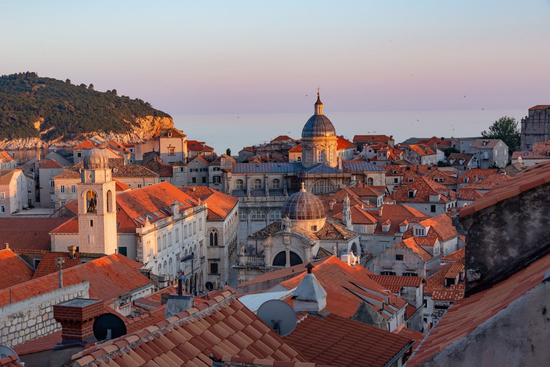 A panoramic view of Dubrovnik's old town at sunset, with historic red-tiled roofs, ancient city walls, and the calm blue Adriatic Sea.