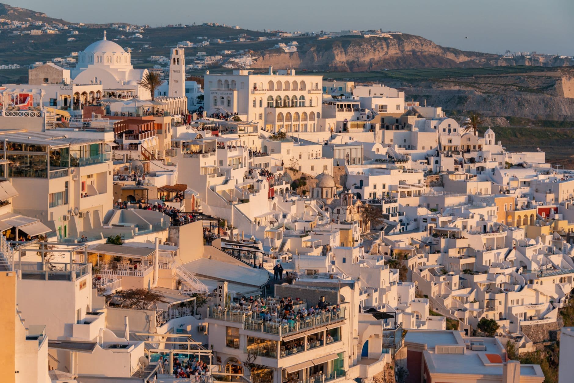 A picturesque sunset scene over the caldera in Santorini, Greece, with iconic white buildings and blue-domed churches bathed in golden light.