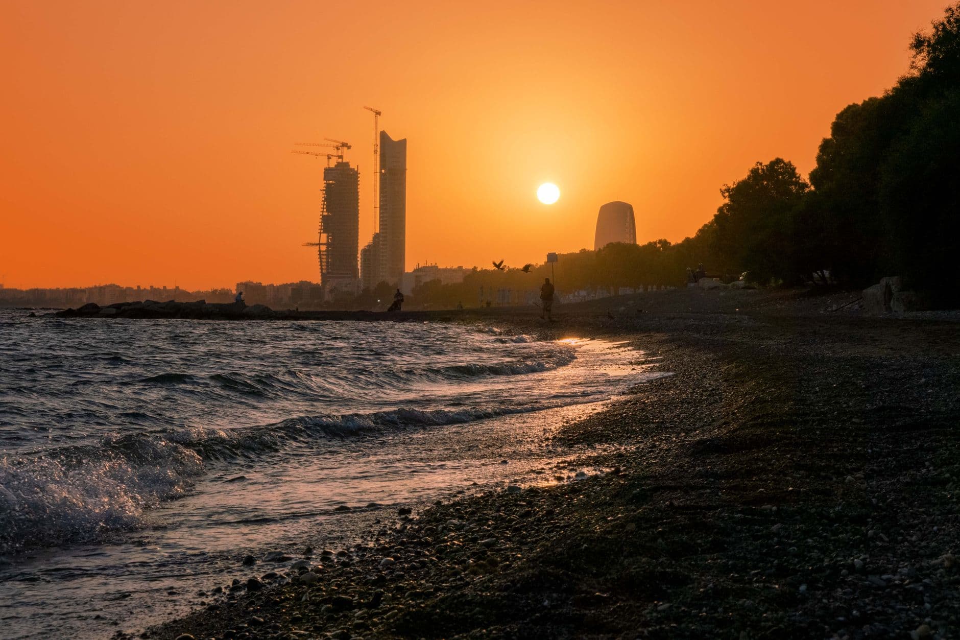 A vibrant sunset over a calm beach in Cyprus, with golden light reflecting on the water and a few distant figures enjoying the view.
