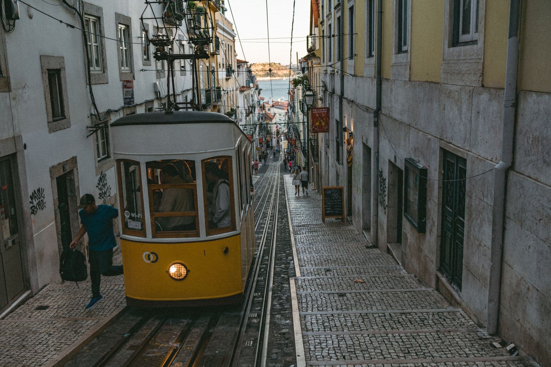 A vibrant yellow tram navigating a narrow, cobbled street in Lisbon's historic Alfama district, with colorful buildings and laundry hanging from balconies.