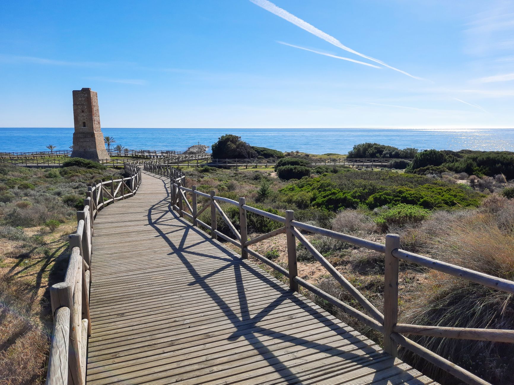 A view of the sandy dunes and wooden boardwalk at Cabopino beach in Marbella, with the Mediterranean Sea in the background.