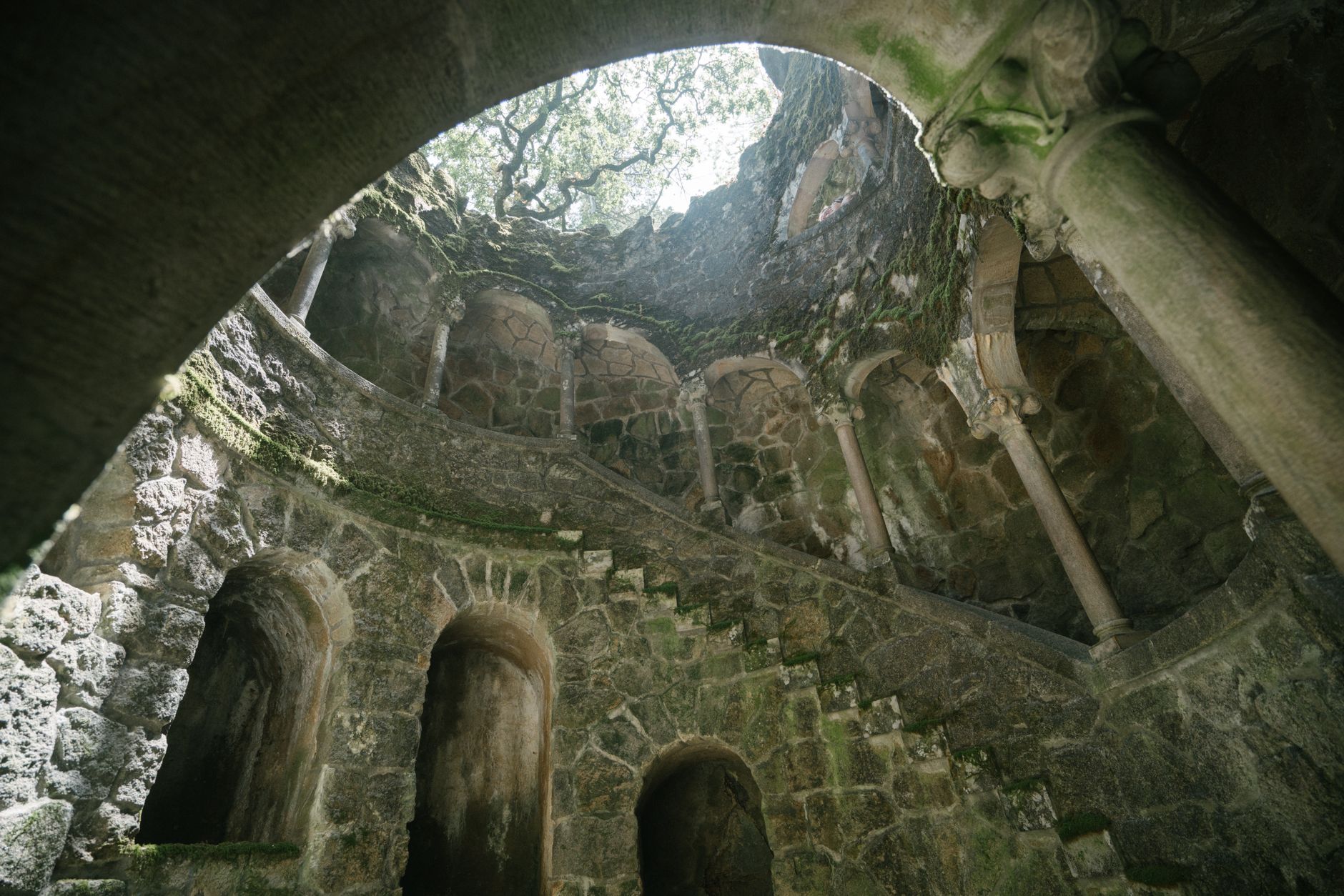 The mysterious spiral staircase of the Initiation Well at Quinta da Regaleira in Sintra, Portugal, with mossy stones and dim lighting.