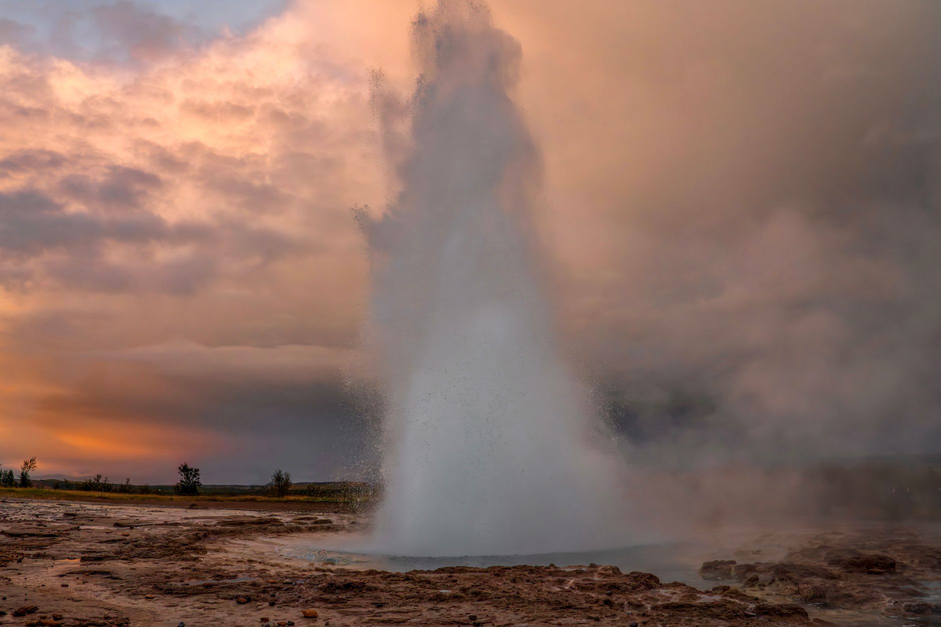 The Strokkur geyser erupting high into the air at the Geysir geothermal area in Iceland, surrounded by steaming earth and distant hills.