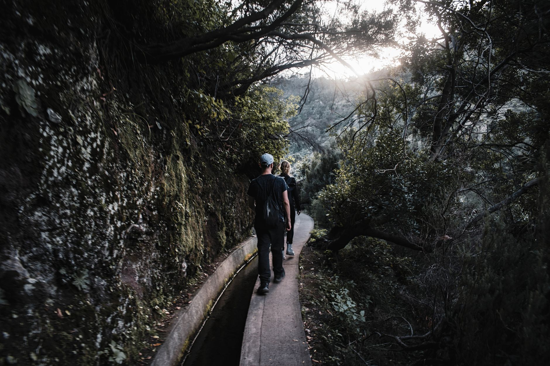 A narrow levada trail winding through lush green vegetation, with a small waterfall cascading into the water channel, on the island of Madeira.