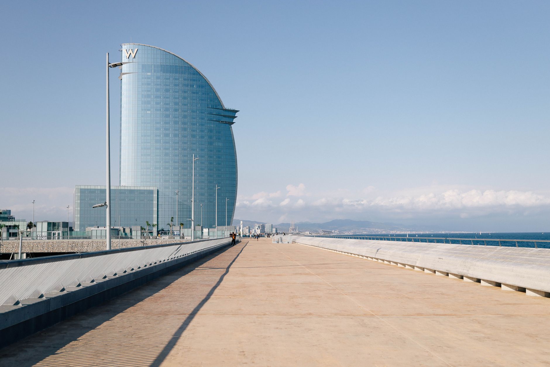 People walking and cycling along the sunny promenade of Barcelona's Barceloneta beach, with the Mediterranean Sea and city skyline in the background.