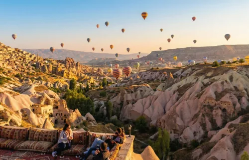 A wide-angle landscape photo captures colorful hot air balloons rising over the rocky formations of Cappadocia at sunrise, with people watching from a terrace—a top destination in a Turkey travel guide