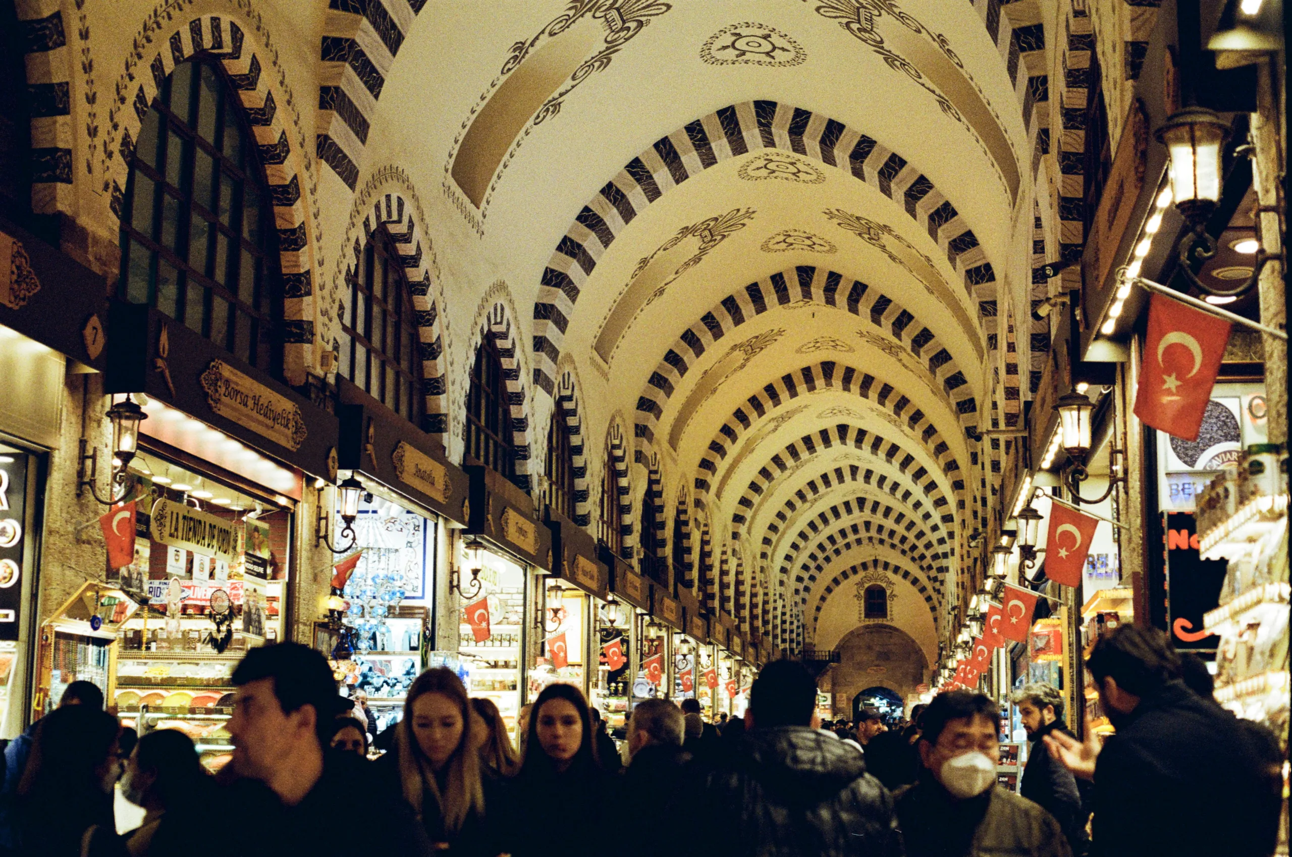 A bustling main corridor inside Istanbul's Grand Bazaar, filled with shoppers, colorful hanging lamps, and historic arched ceilings.