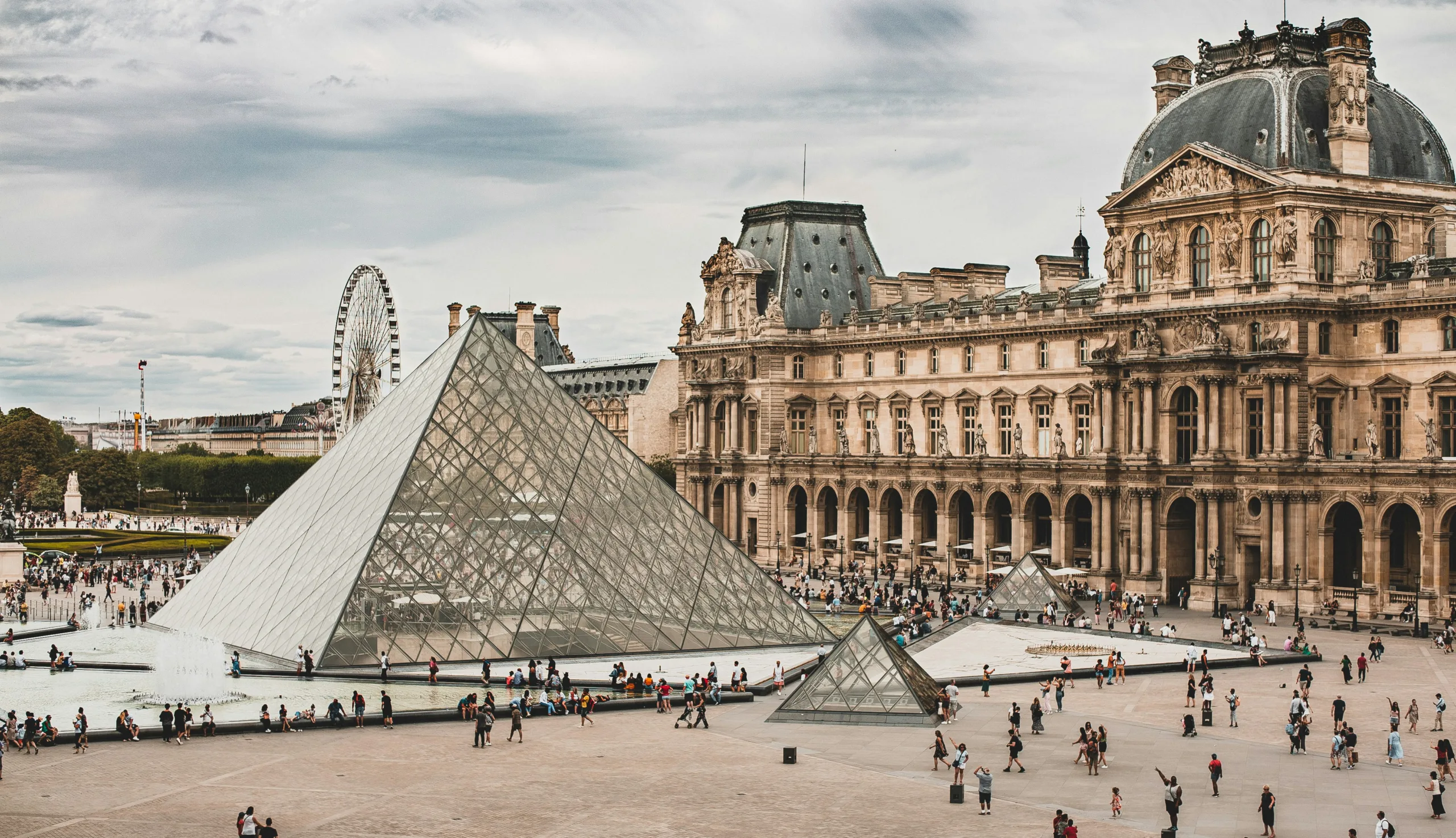 A bustling daytime view of the Louvre Museum courtyard in Paris, with crowds of visitors gathered around the iconic I.M. Pei glass pyramid.